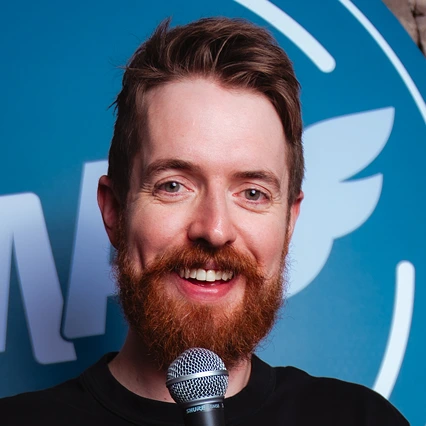 Scottish comedian Rosco McClelland smiles while holding a microphone in front of an LMAOF backdrop.