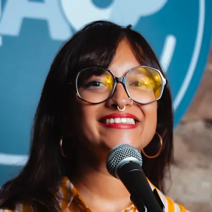 Comedian Shalaka Kurup smiles behind a microphone while wearing oversized glasses in front of an LMAOF backdrop.